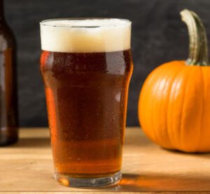 Pint of amber beer with a foamy head on a wooden surface, next to a small orange pumpkin.