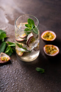 Glass of sparkling drink with ice, passion fruit slices and mint leaves on a dark tabletop; halved passion fruits nearby.