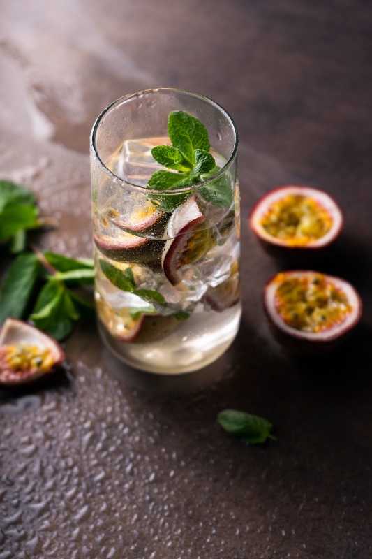 Glass of sparkling drink with ice, passion fruit slices and mint leaves on a dark tabletop; halved passion fruits nearby.