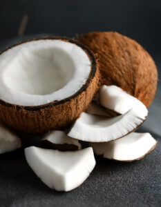Halved coconut with white flesh and several chunks scattered on a dark surface on a black background.