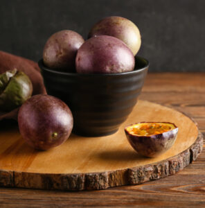 Purple passion fruits piled in a black bowl on a wooden board, with one cut open showing orange pulp and seeds.