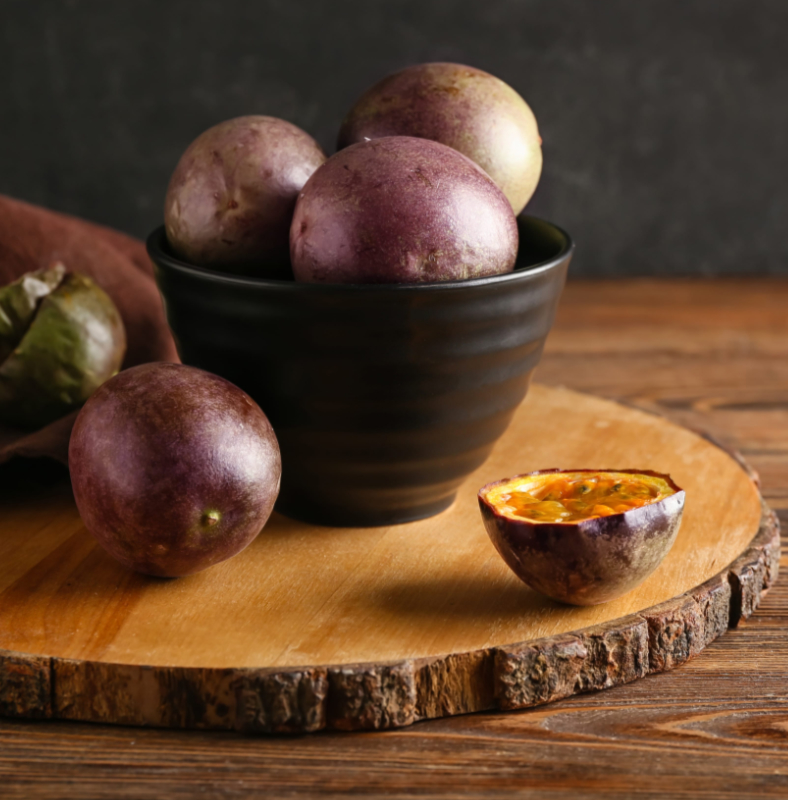Purple passion fruits piled in a black bowl on a wooden board, with one cut open showing orange pulp and seeds.