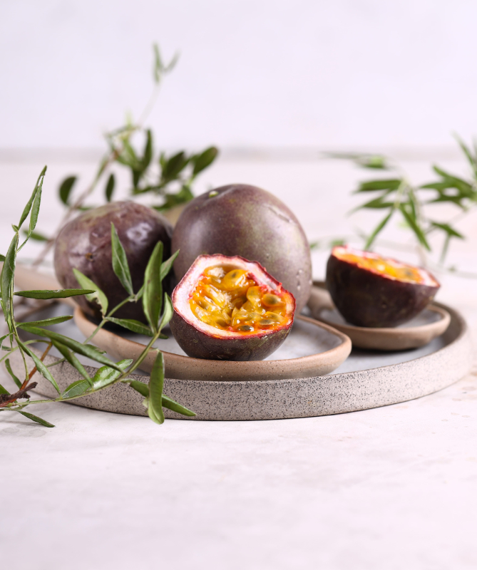 Open passion fruit on a light plate, revealing bright orange pulp and many seeds with green foliage in the background.