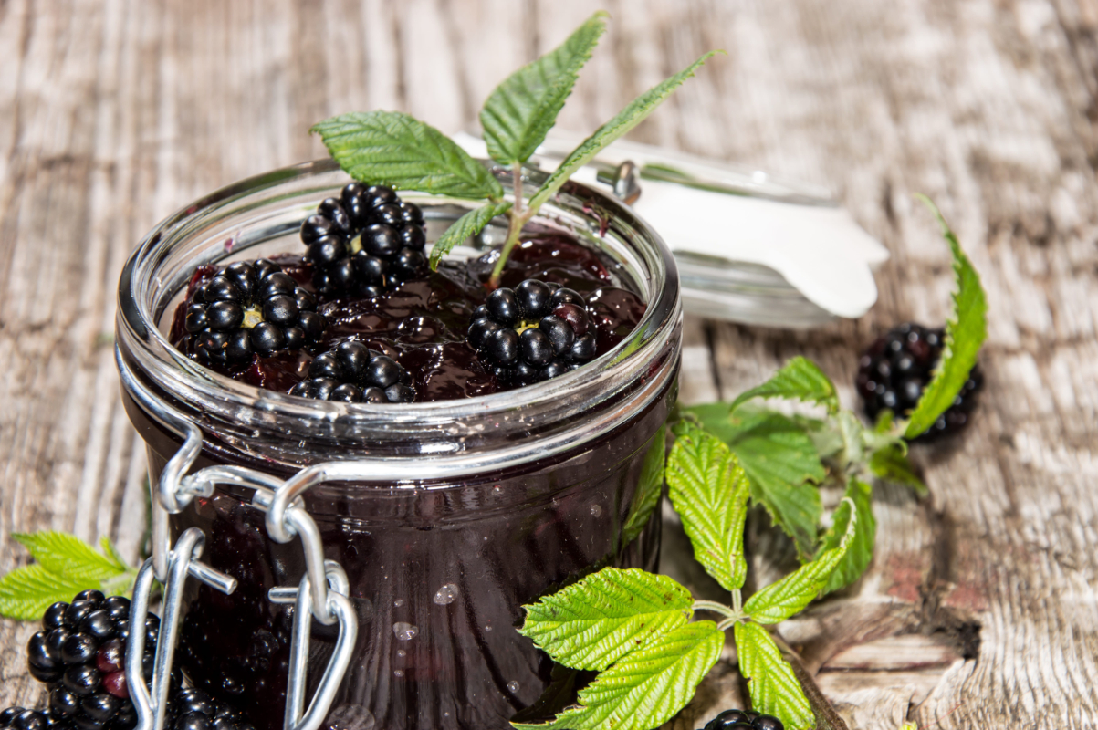 Blackberry Jam in a glass on wooden background | Puree Arete Glass jar of blackberry jam with fresh berries and mint leaves on a rustic wooden table in the background of a kitchen scene