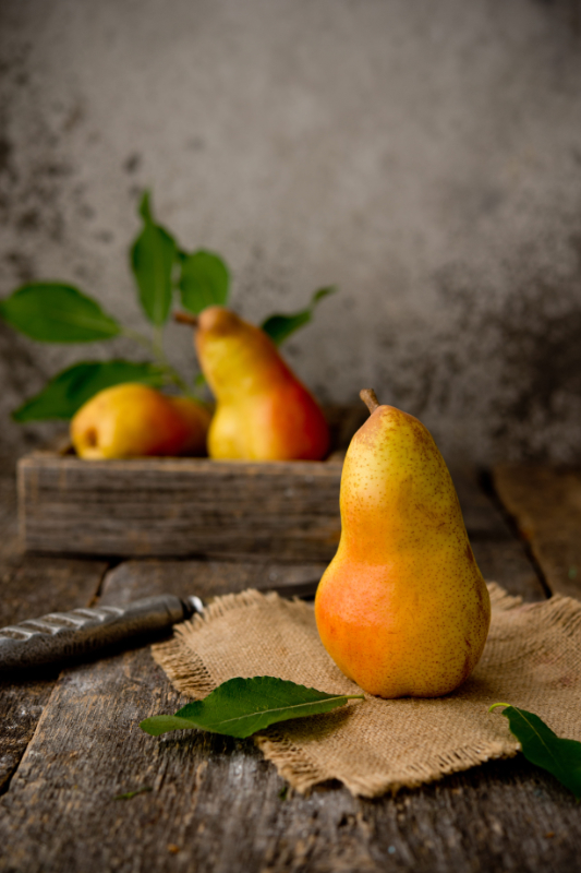Fresh juicy pear on rustic wooden background | Purée Arêté A ripe yellow-orange pear sits on a burlap cloth on a weathered wooden table, with a wooden crate of pears and green leaves blurred in the background.